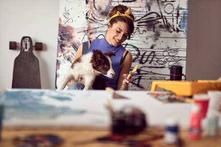 A young female artist is playing with her dog while working on a new painting in a pleasant atmosphere in the studio. Art, painting, friendship, studioの写真素材