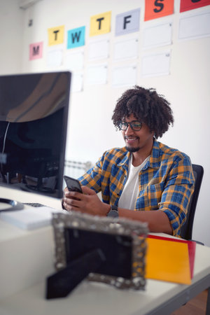 A young man sitting at the desk in a pleasant atmosphere in the office and texting on the smartphone. Employees, job, officeの写真素材