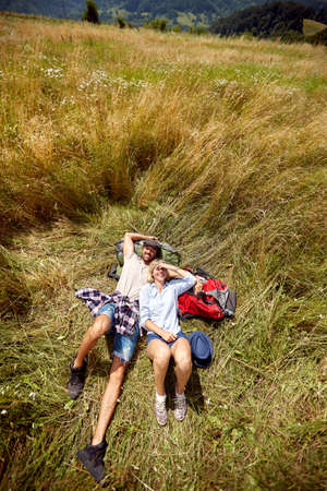 A young couple is relaxing on the grass while walking a meadow on a beautiful sunny day. Hiking, nature, relationship, togetherの写真素材