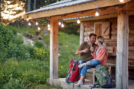 A young couple having chatting and having a drink at the cottage porch in the forest on a beautiful day. Vacation, nature, cottage, relationshipの写真素材