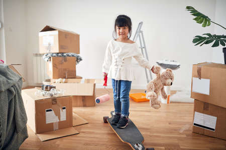 cute little asian girl smiling, standing on boosted board, holding teddy bear and paint roller, surrounded with unpacked cardboard boxes in new apartment.の写真素材
