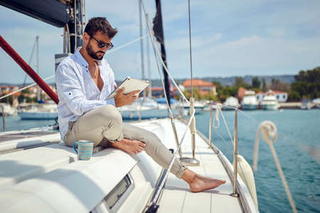 A young man watching a tablet content while sitting on a yacht and riding through the dock on a...