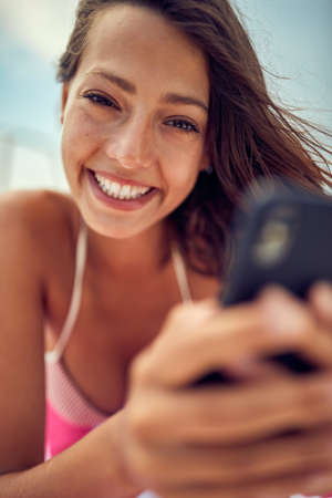 Close-up of a young sexy girl using a smartphone while posing for a photo and  enjoying a sunbath on a beautiful summer day. Summer, sea, vacationの写真素材