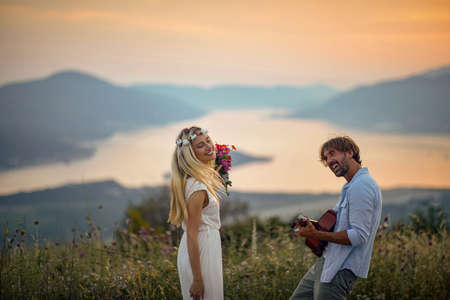 A young groom is playing a guitar for his bride on a beautiful sunset in the nature. Relationship,...