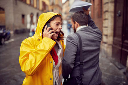 A young woman who is walking the city in a hurry manner and speaking on the phone on a rainy day hit...