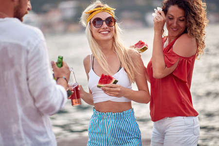 A group of friends having fun on the dock on the seaside while having a drink and juicy watermelon on a beautiful day. Friendship, holiday, seaの写真素材