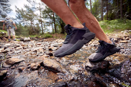 Close-up of man's legs while crossing a creek during a hiking on a beautiful sunny day. Hiking,...