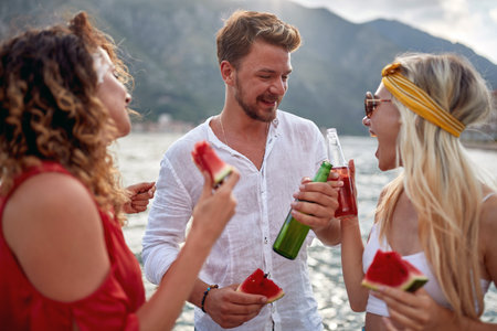 Happy people enjoying a day at the lake laughing and eating watermelon and drinking beers.の写真素材