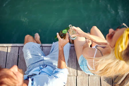 Bird-eye view on a young happy couple sitting on the dock on the seaside and chatting with drink on...
