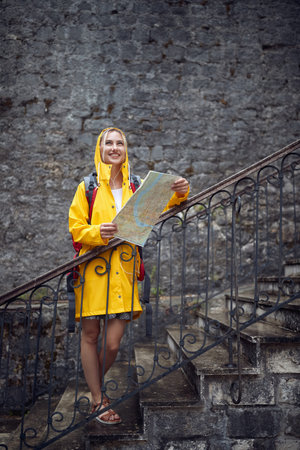 Woman in raincoat holding map, feeling joyful. Summertime rain. Travel, city, lifestyle concept.