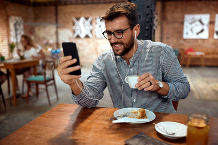 Young handsome man sitting in cafe, enjoying coffee and looking at smartphone. Video call, chatting, listening to music, scrolling.の写真素材