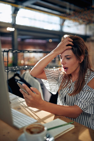 Surprised young woman looking at smarthone sitting at table in front of desktop computer. Business, student, employee concept.の写真素材
