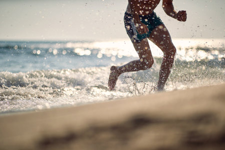 Young athletic man on beach running in water. Focus on lower part of body. Wellness, lifestyle,...