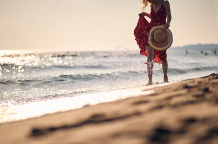 Young woman in waving red summer dress walking on beach holding fashionable straw hat. Vacation, holiday, lifestyle concept.の写真素材