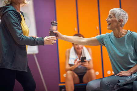 Senior woman in sportswear offering her friend a water bottle. Group of women in gym locker room...
