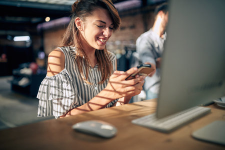 Beautiful young woman sitting at table in front of desktop computer using smartphone, feeling joyful. Business, student, office job concept.の写真素材