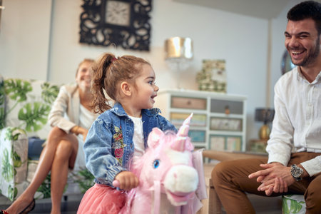 Young family is captured in a moment of pure joy and happiness. The mother can be seen laughing in the background, while the father stands next to their adorable toddler daughter who is riding a rocking horse toy. The trio is having a great time as they bond and create precious memories together.の写真素材