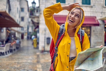 Walk in city in rain. Young tourist woman in raincoat with map, feeling happy, smiling. Travel in new city. Tourism, lifestyle, fun, concept.の写真素材