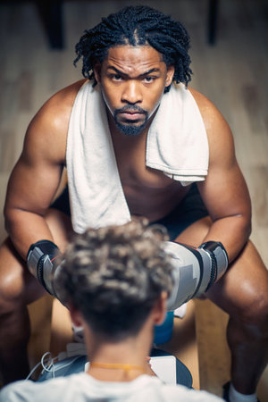 High agle shot of strong muscular young shirtless man wearing gloves, sitting in front of trainer,...
