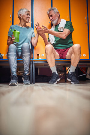 Senior man and woman sitting in gym locker room, talking and sharing an apple. Helath, active lifestyle, senior life concept.の写真素材