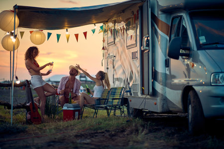 Three friends toasting and having fun outdoors, in front of camper rv. Summertime sunset. Travel, holiday, togetherness, lifestyle concept.の写真素材