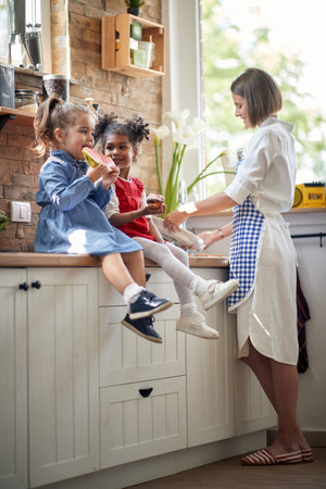 Two cute little girls sitting on countertop eating a snack, watermelon and muffin, by their mother washing dishes. Home, family, lifestyle concept.の写真素材