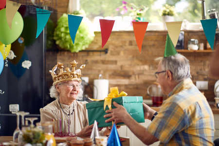 Senior couple celebrating together, elderly birthday woman recieving birthday gift present from her husband sitting at dinner table at home. Celebration, senior life, lifestyle concept.の写真素材