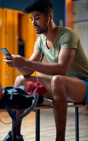 Handsome young sporty man with earphones and smartphone sitting in gym locker room, getting ready for workout session. Health, lifestyle, wellness concept.の写真素材