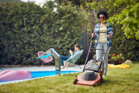 Peaceful summer day, where a woman diligently tends to the lawn while her husband finds respite and relaxation in the comforting embrace of a poolside chair.の写真素材
