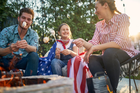 Family gathers in their backyard, basking in the joy of togetherness. The parents and their daughter are seen indulging in the simple pleasure of roasting marshmallows over a crackling fire.の写真素材