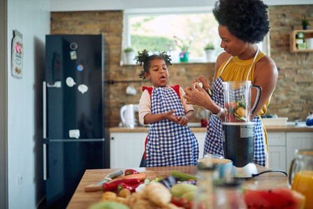 Young mother in the home kitchen with her little daughter, cutting up vegetables and tasting cucumber slices, girl acting silly. Home, family, lifestyle concept.の写真素材