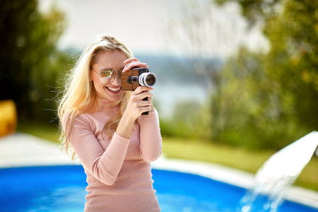 Young joyful blonde woman holding video camera and recording outdoors by the pool in nature, feeling...