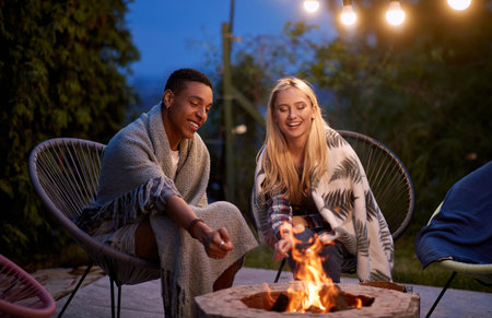 multiracial adult young couple, roasting marshmallow, smiling, sitting by the fire covered with...