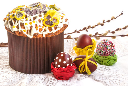 Easter composition: eggs, typical cake (covered with icing, sprinkled with color pops and candied flowers) on lace napkin and willow branches in background. Selective focus, white background, horizontal.の写真素材