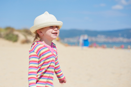 Little two year old girl with blue eyes in a bright wicker hat, standing on the beach near the seaの写真素材