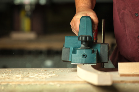 Carpenter working with electric planer on wooden plank in workshop.