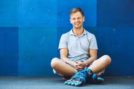Portrait of happy handsome young roller in inline skates sitting against blue wall.