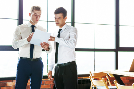 Two young business men discussing working moments standing against bright window.の写真素材