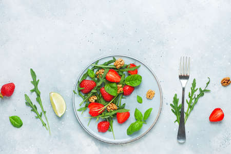 Summer strawberry salad. Fresh arugula, basil, sliced strawberries, nuts with lime on grey stone background. Healthy vegan food concept. Flat lay, top view, copy spaceの写真素材
