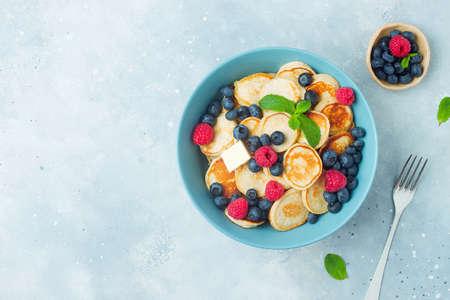 Trendy food - mini pancakes cereal. Heap of mini cereal pancakes with summer fruits in bowl on grey blue background. Copy space. Top view or flat lay.の写真素材