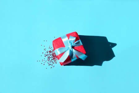 Red gift and confetti with hard shadows on blue background. Christmas, new year, holiday concept. Trendy style. Minimal, flat lay, top view, copy spaceの写真素材