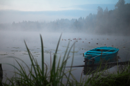 Lonely boat on a peaceful lake. Foggy autumn sunrise twilight. Bright blue turquoise shallop quiet forest, swamp plants, smoke mist on still calm water. Russia Moscow area nature landscapeの写真素材
