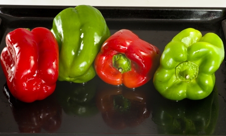 Four peppers, two red and two green, soaked in oil, ready to be tucked into the oven and roast  They are on a black tray with a few drops of oilの写真素材