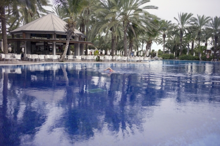 It is a very blue water pool  The Palm trees are reflected in the water  There is a person with a red hat, swimming  On the other side of the pool there is a place for drinks and chairsのeditorial素材