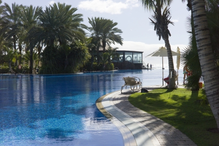 A very blue pool, with palm trees in the garden, loungers and parasols can be seen  There are some people  You can see the sea in the backgroundのeditorial素材