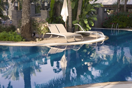 A very blue pool, with Palm trees in the garden, loungers and parasols can be seen  The reflections of the Palm trees can be seen in the waterのeditorial素材