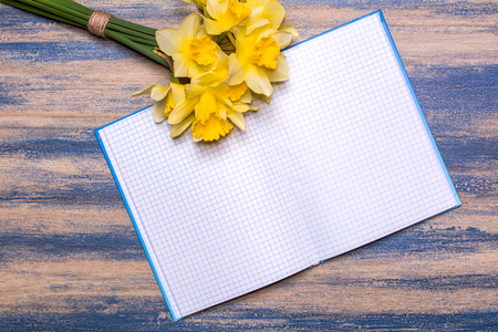 Notepad on a wooden board. Yellow daffodils flowers on a wooden table. Bloomed blue paint. Background with space for text. View from above, flat lay design.の写真素材