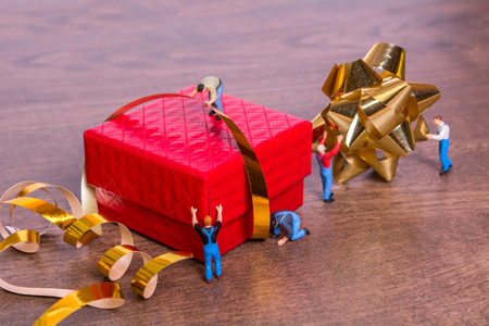 Miniature people and a gift box on a wooden background. The process of packing gifts. Gold ribbon, bow, festive beads. Creative concept of teamwork.の写真素材