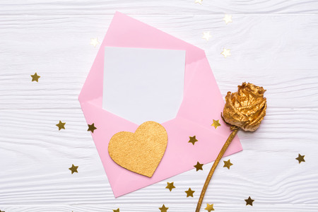 Flat lay pink envelope with a note, gold rose and gold heart on a white wooden background. Asterisks of confetti. Romantic composition. View from above.の写真素材