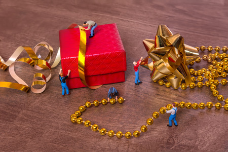 Creative concept with miniature people and a gift box on a wooden background. The process of packing gifts. Gold ribbon, bow, festive beads.の写真素材